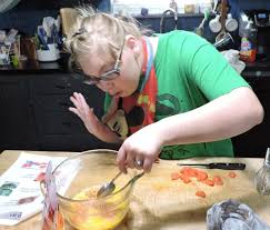 una joven se encuentra mezclando con su mano izquierda, huevos con una cuchara en un recipiente de vidrio, la joven utiliza anteojos.