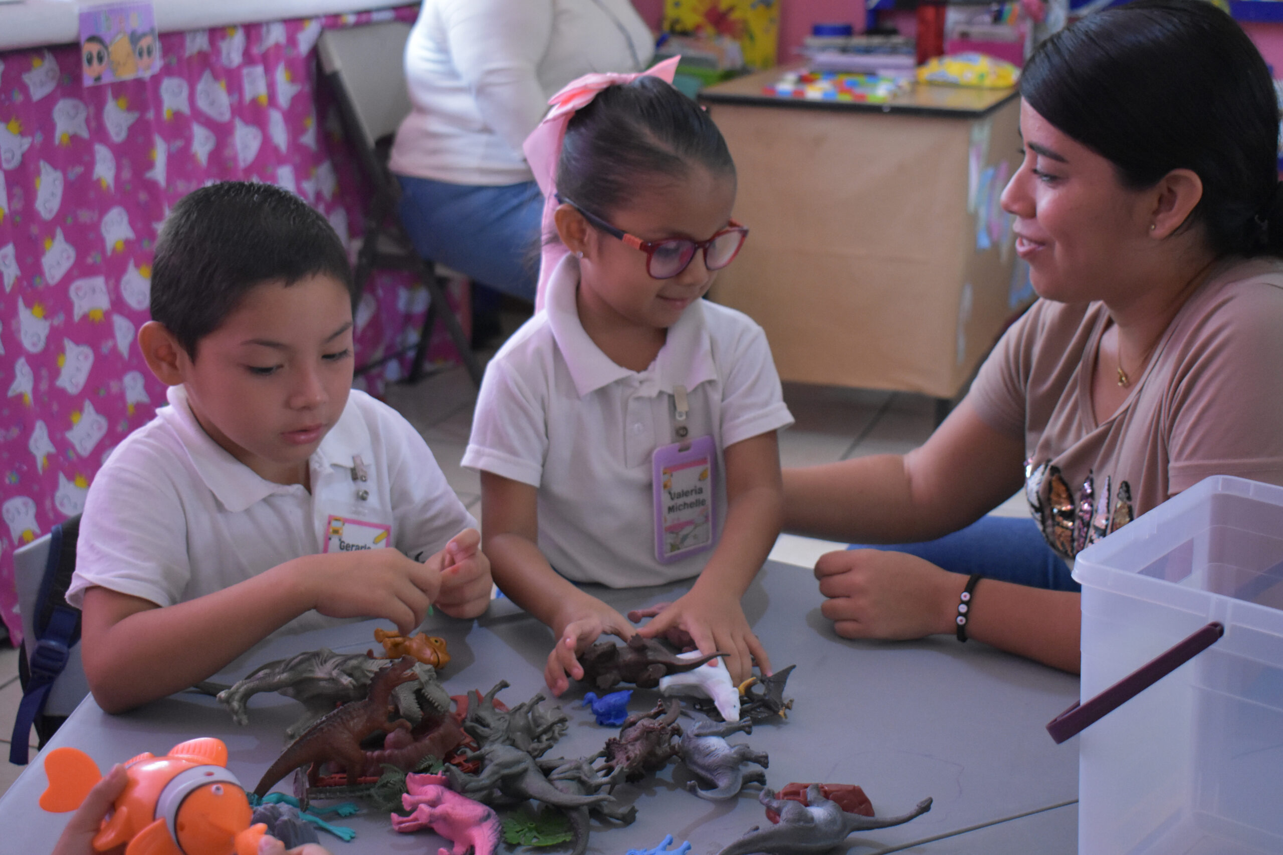 Dos niños pequeños juegan con dinosaurios de juguete en una mesa mientras una mujer está sentada a su lado, sonriendo y charlando con ellos. El fondo incluye decoraciones coloridas y una persona sentada en otra mesa.