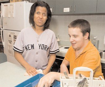 Una mujer con jersey neoyorquino sonríe junto a un hombre con camiseta naranja que carga o descarga un lavavajillas en una cocina.