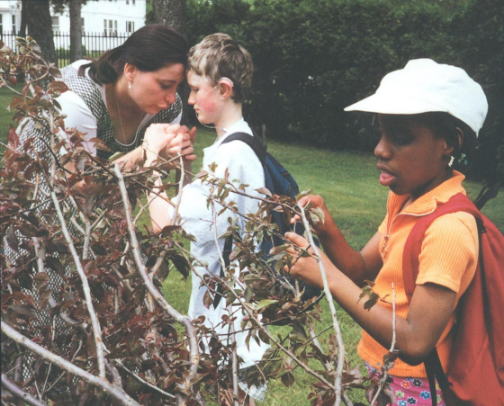 Una mujer coge y besa la mano de un niño, mientras una niña con camiseta naranja y gorro blanco examina unas ramas cercanas. Los tres están al aire libre en una zona de césped.