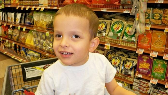 Un joven de pelo castaño corto y camiseta blanca está sentado en un carrito de la compra dentro del pasillo de un supermercado, con estantes de comida envasada detrás de él.
