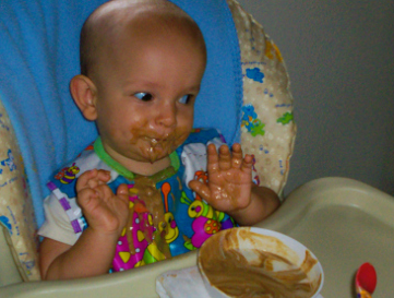 Una bebé sentado en una trona con comida embadurnada en la cara, las manos y el babero, mirando a un lado con expresión juguetona. Un cuenco de comida desordenada y una cuchara están en la bandeja frente a ella.