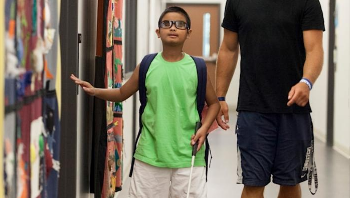 Un chico joven con gafas y un bastón blanco camina por un pasillo con un hombre a su lado. El chico toca la pared con una mano y lleva una mochila. El hombre lleva una camisa negra y pantalones cortos.