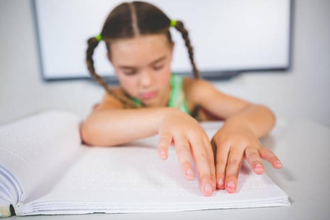 Una niña con trenzas y blusa verde se inclina sobre un libro en braille, usando sus manos para leer.