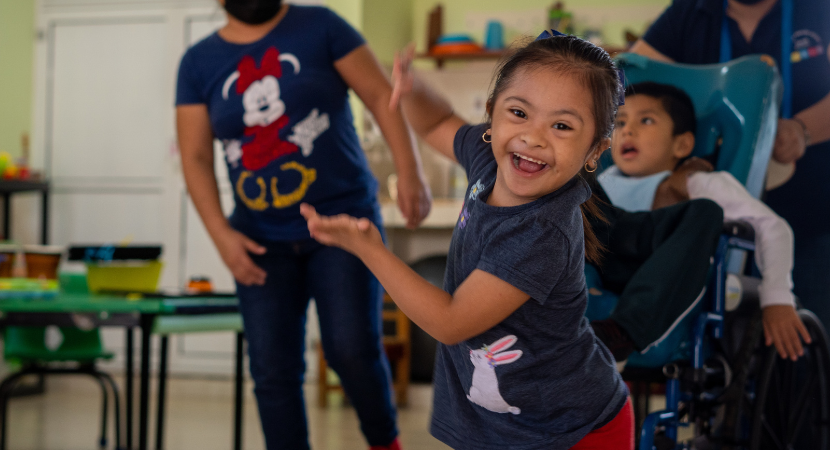 Una niña con camiseta azul y pantalones rojos sonríe a la cámara y gesticula con sus manos. Al fondo, es posible ver a un adulto y a otro niño en su silla de ruedas.