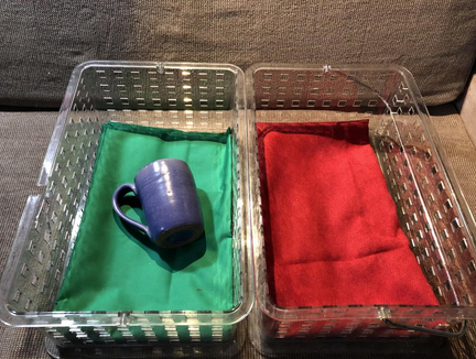 Two bins. One has a cup laying on a green kitchen cloth and the other one has only a red kitchen cloth,