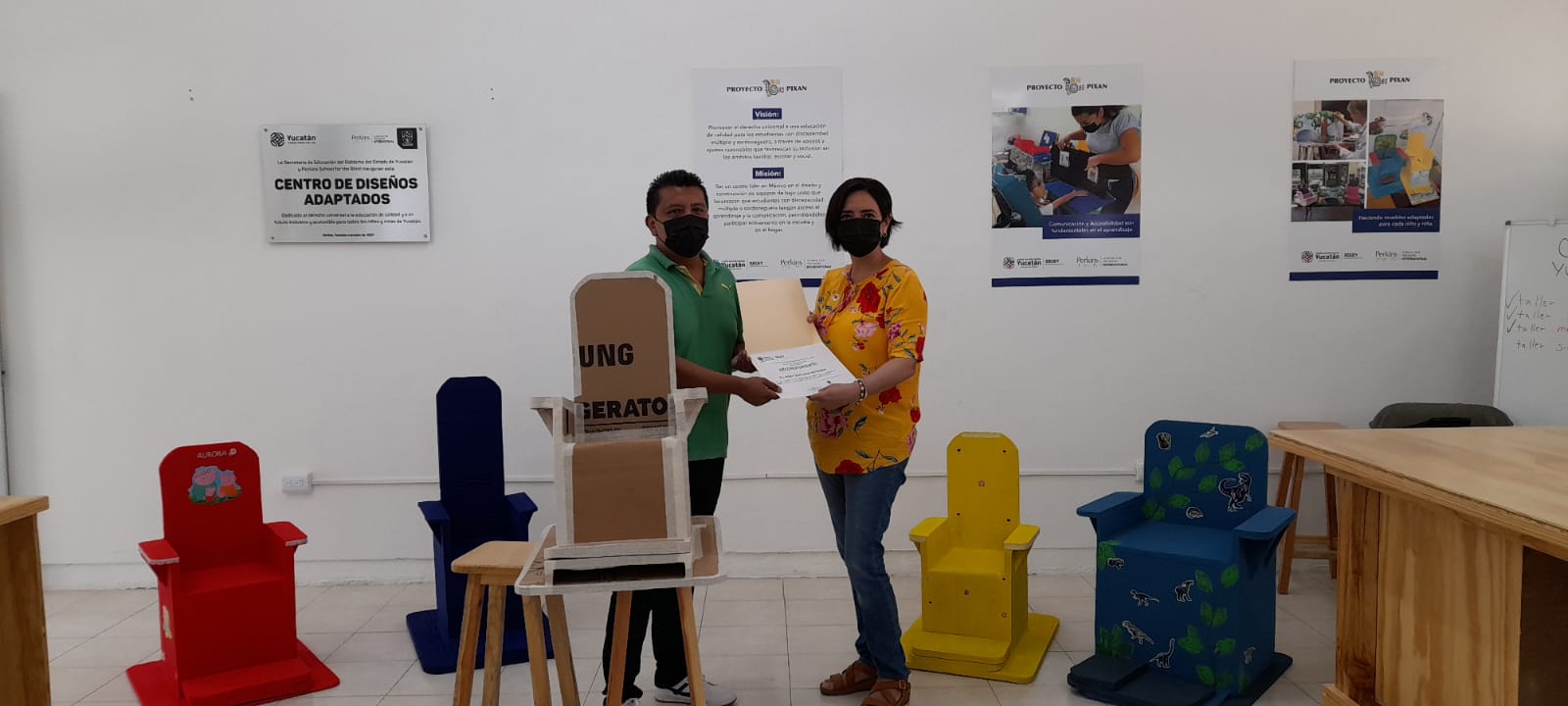 Imagen: Un hombre y una mujer sostienen un reconocimiento. Se ven al fondo5 sillas acabadas hechas con cartón . 4 pintadas de colores y una sin pintar