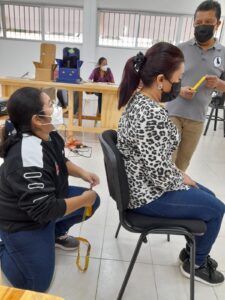 Imagen: Dos mujeres una sentada la otra detrás con una cinta métrica escuchan a un jóven que da indicaciones