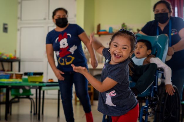 Tere, una niña bailando y sorreindo en su clase. Ella leva pantallones rojo y una camiseta azul.