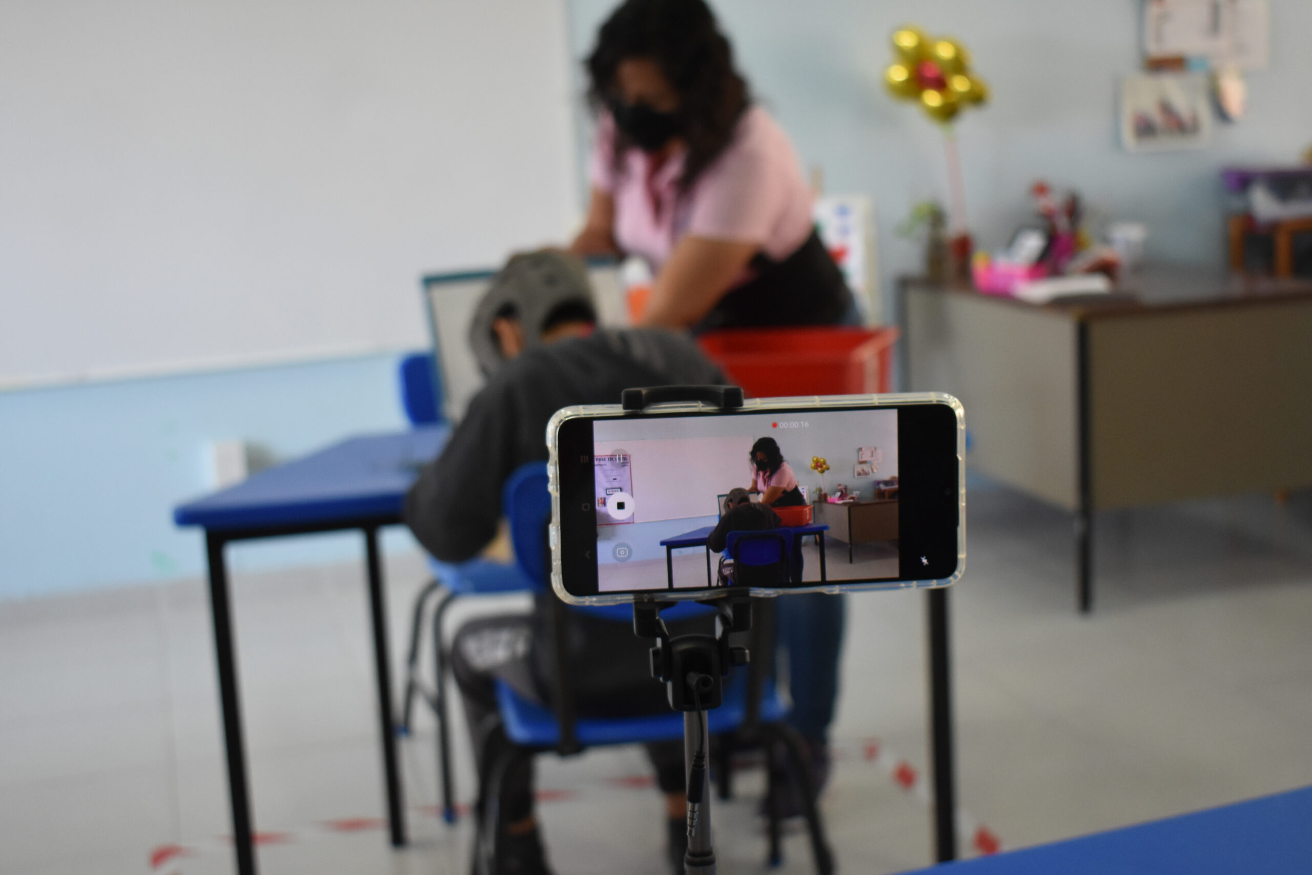Imagen: En primer plano un celular grabando escena, en segundo plano una maestra enfrente de un estudiante con discapacidad de quien sólo se ve la espalda, ambos trabajan con materiales en la mesa.