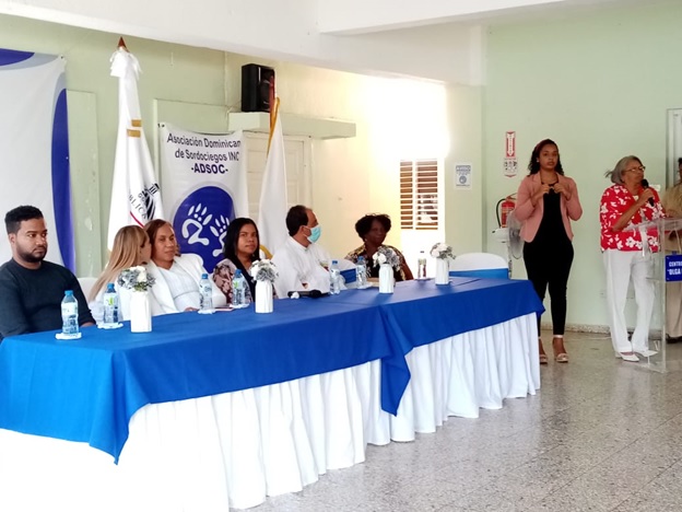 Seis personas sentadas en la mesa del presidium, una mujer habla frente al microfono mientras otra mujer interpreta con senas