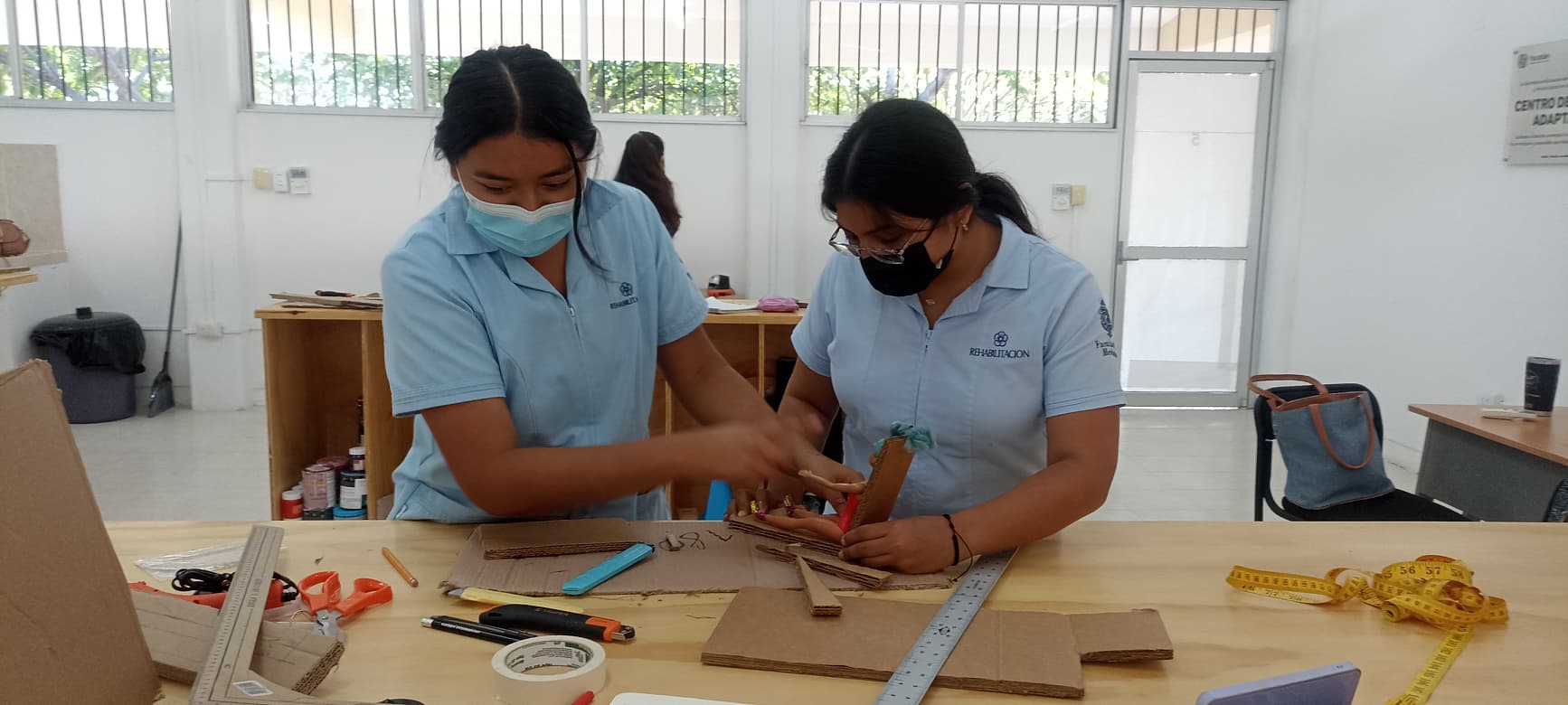 Dos estudiantes de rehabilitación de la Universidad Autónoma de Yucatán cortan cartón y miden una silla de cartón para una muñeca
