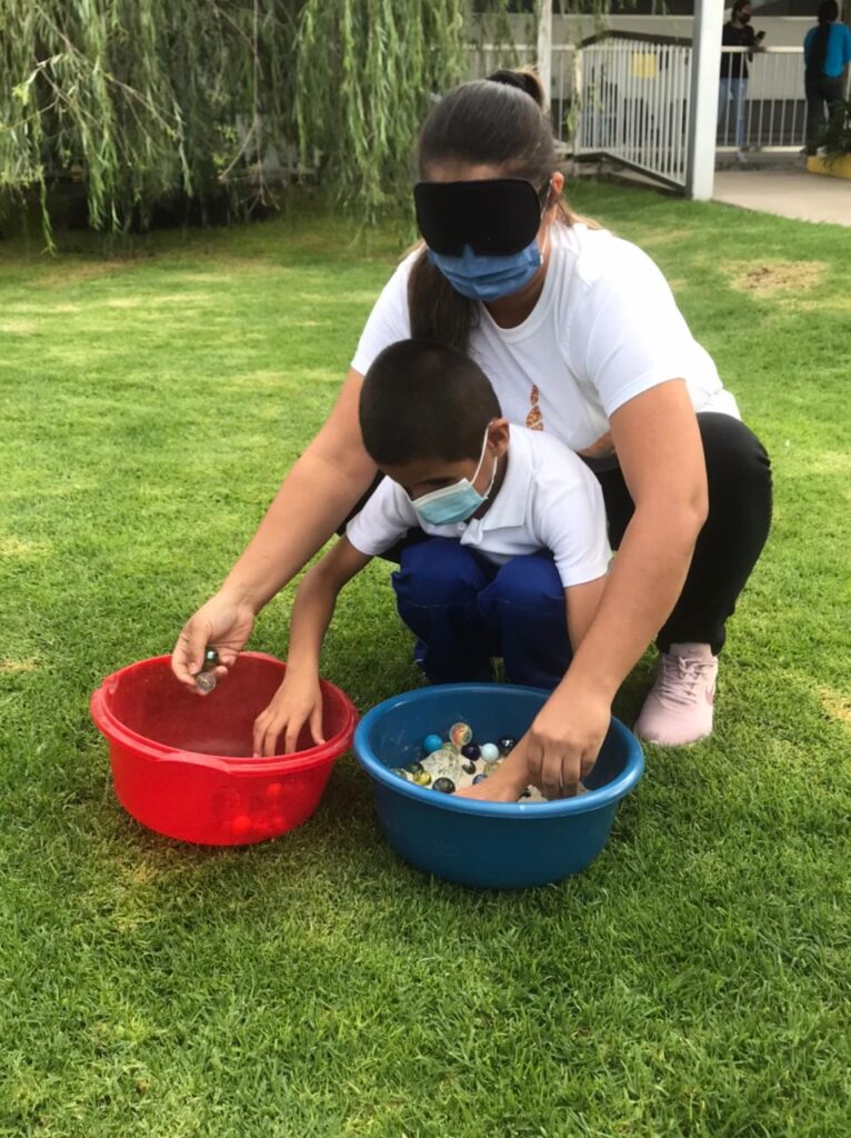 Madre e hijo están en cuclillas en el pasto jugando con unas pelotas que se encuentran dentro de unas bandejas, la madre tiene los ojos vendados, el niño tiene discapacidad visual