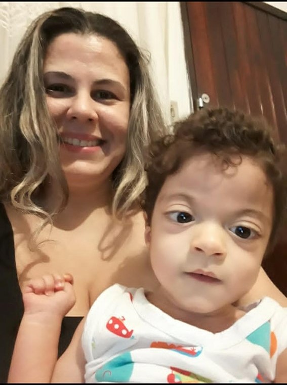 Suzana sorrindo para a camêra e segura seu filho Pedro, que olha atentamente para a frente.