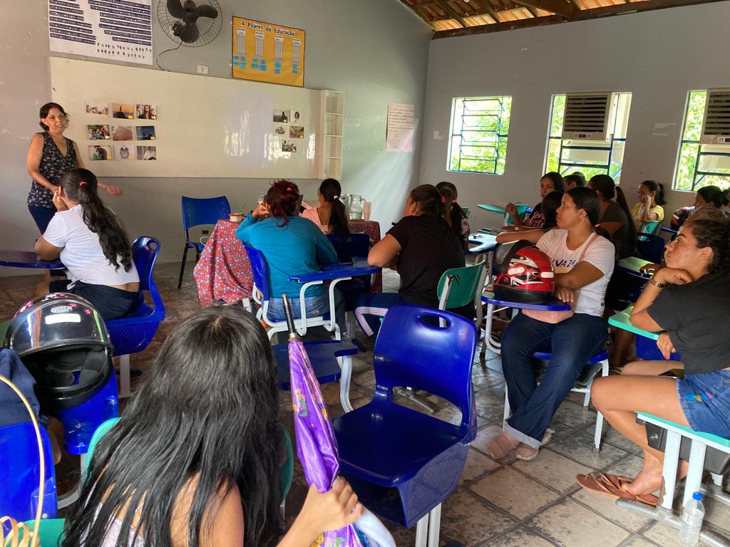 Uma sala de aula com quadro branco e Lidiane na frente vestindo jeans e blusa  florida, falando para uma sala cheia de pessoas sentadas em carteiras escolares azul.