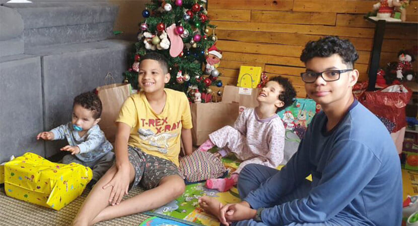 Um fundo com sofá cinza, painéis de madeira e uma árvore de Natal com presentes. Em frente à árvore na esquerda, Lucas tentando alcançar um pacote amarelo, Arthur sorrindo, Micaela de pijama rosa sorrindo e Arhur de pijama azul olhando diretamente para a câmera.