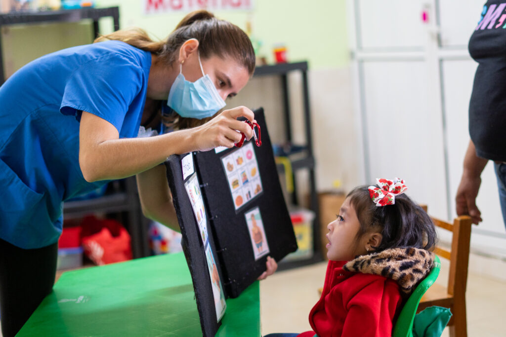 Una maestra vestida de azul y con una máscara facial sostiene un libro de comunicación que muestra imágenes a una niña frente a ella. La niña se sienta en una silla verde, usa un abrigo rojo y mira directamente al libro. Están en un salón de clases, el fondo esta borroso