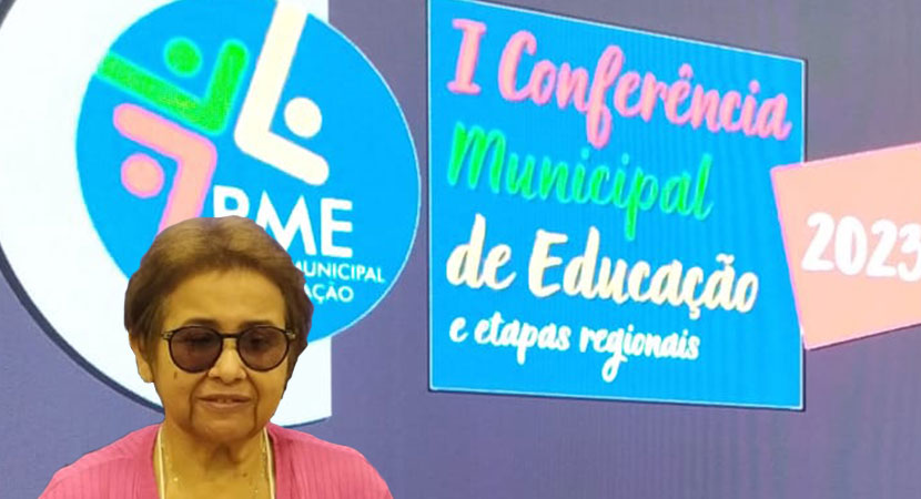 Eulalia em frente a um painel da Primeira Conferencia Municipal de Educação. Ela veste um conjunto preto e rosa.