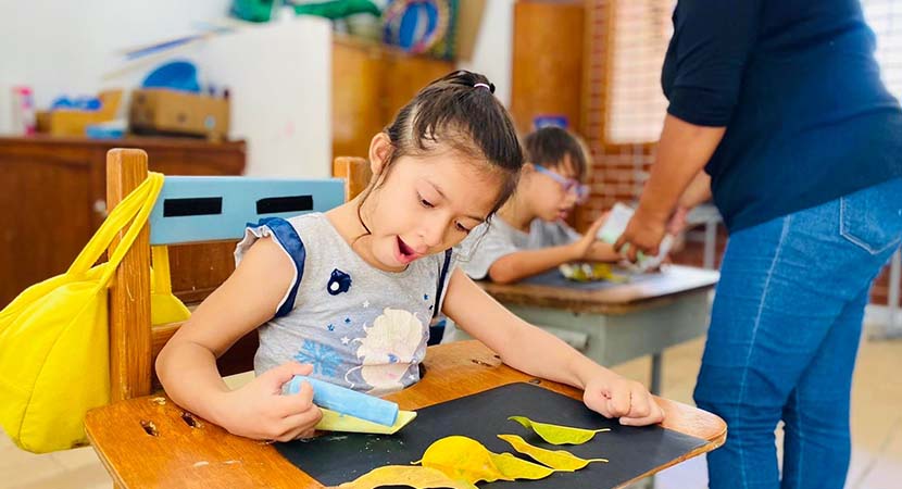 Una niña en un salón de clases trabajando en una hoja de papel negro con collages amarillos.