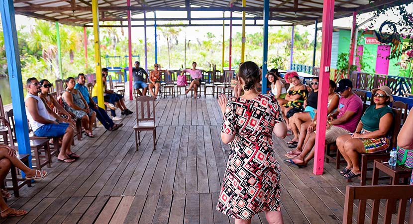 Lidiane de costas para a camera, com um vestido colorido e descalça. A sua volta, um grupo de familias sentados observam a sua fala. 