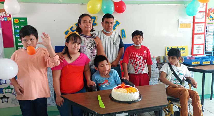 Foto de Adonis con sus compañeros y maestra en frente de un pastel de cumpleaños.