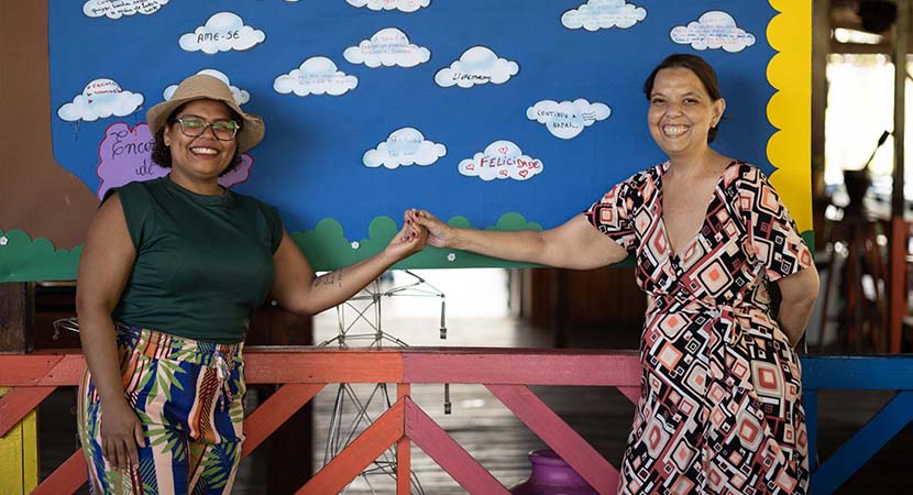 Lidiane e outra mãe, estão em pé em frente a um mural escolar colorido. As duas esrendem as mãos entre elas.