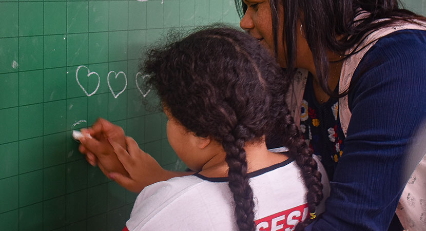 Menina escrevendo na pizzarra com a ajuda da professora
