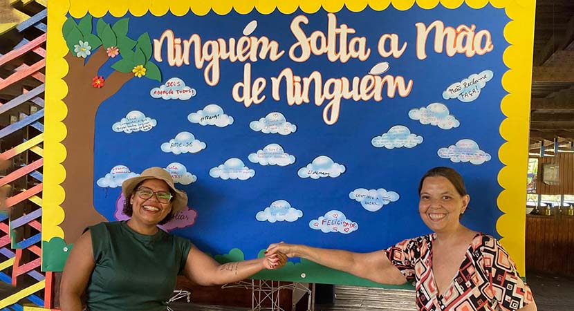 Lidiane, madre y coordinadora familiar de Perkins en Brasil, y Kelly Cristina, otra madre, están paradas frente a un colorido mural escolar que dice: "Nadie suelta la mano de nadie". Las dos extienden los brazos y se toman de la mano.