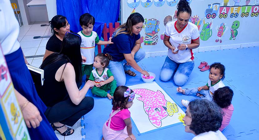 Un grupo de cinco niños pequeños y cuatro adultos en un salón de clases de un programa de atención temprana, sentados sobre una alfombra azul y mirando la imagen de un delfín rosado.