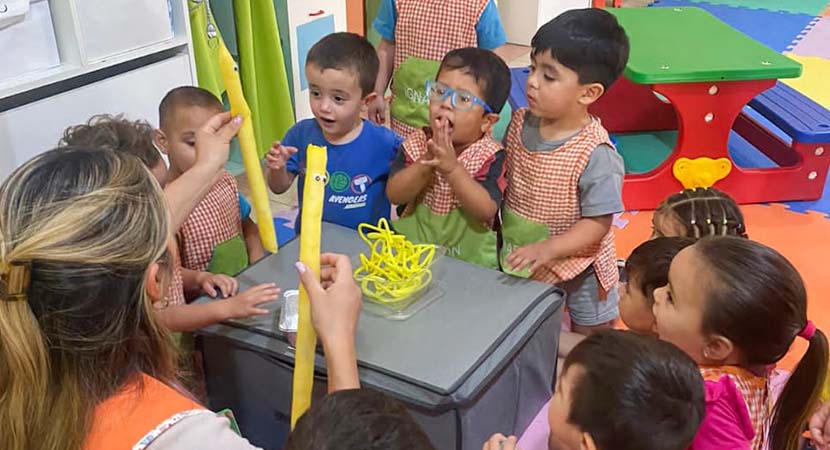 Un grupo de 12 niños pequeños está sentado alrededor de una caja. En frente, la maestra también está sentada con algunos materiales amarillos, contando una historia.