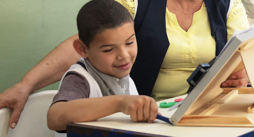 Un niño y una maestra en un salón de clase sentados y mirando a un atril.

