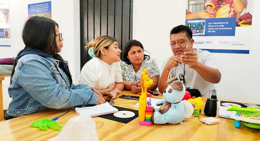 Tres mujeres y un hombre sentados en una mesa larga de madera. En la mesa hay algunos juguetes coloridos y otros materiales. El hombre demuestra a las participantes un cable.
