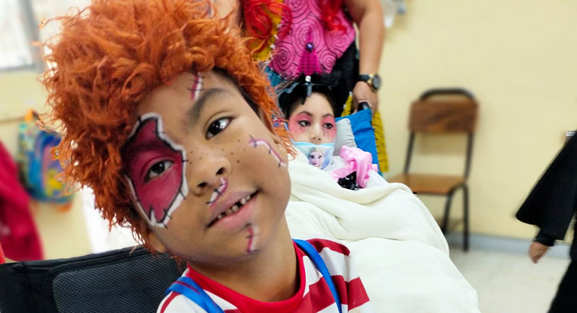 Un niño en su silla de ruedas, vestido con un disfraz en los colores rojo y azul, mirando y sonriendo a la cámara.