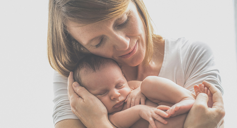 Una mujer con los ojos cerrados vistiendo una camisa blanca, sostiene a un pequeño y somnoliento bebé en sus brazos