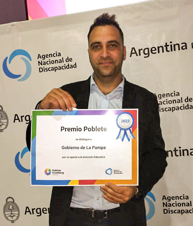 Dr. Ladio posando con su certificado de reconocimiento. Se encuentra frente a un fondo blanco que muestra diferentes logotipos gubernamentales. Ladio viste un traje oscuro y una camisa clara