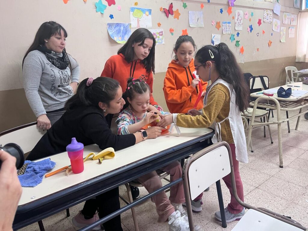 En un salón de clases, Vicky está sentada en un pupitre tocando un objeto. A su alrededor, hay tres de sus compañeros interactuando con ella, y dos maestros. La pared está llena de dibujos coloridos. Sobre la mesa, hay una botella de agua rosa y una cáscara de plátano.