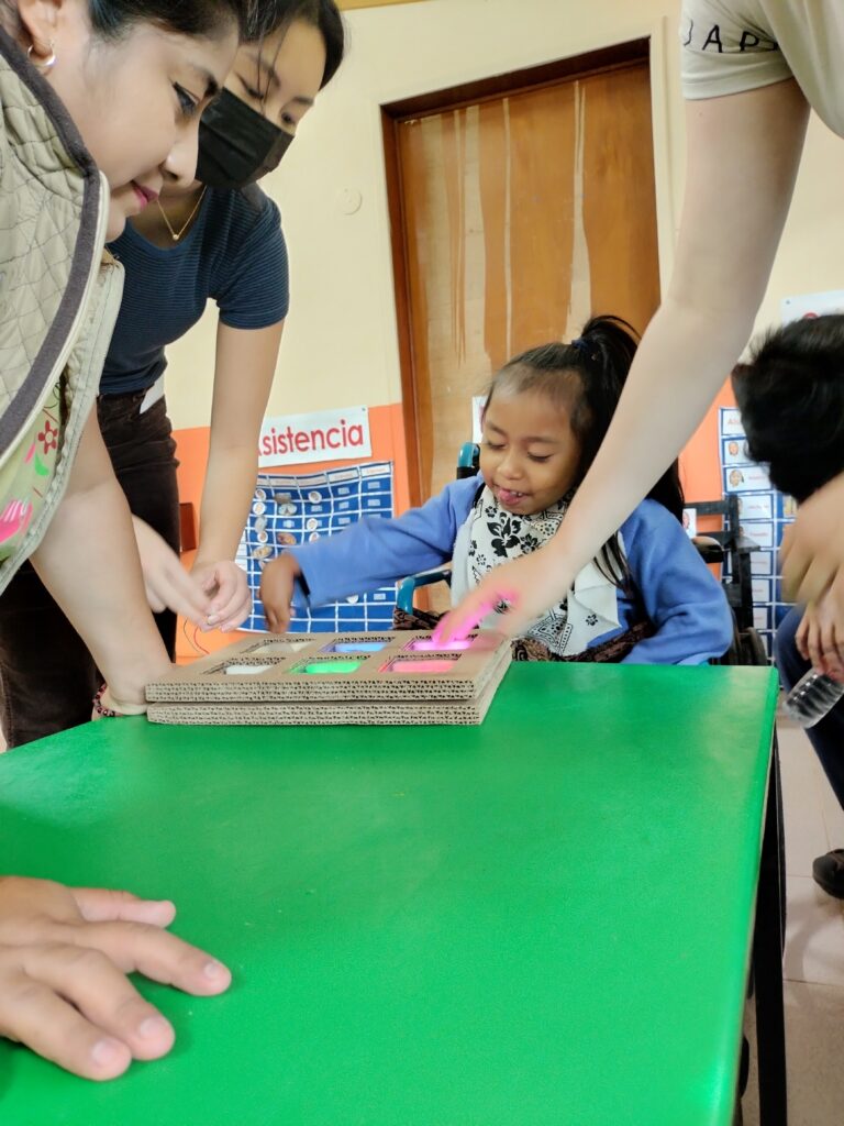 Una niña sentada en una silla de ruedas alcanza una mesa verde donde encuentra una tableta electrónicamente alimentada hecha de cartón. A su alrededor, tres adultos observan su emoción al ver las luces en la tabla.