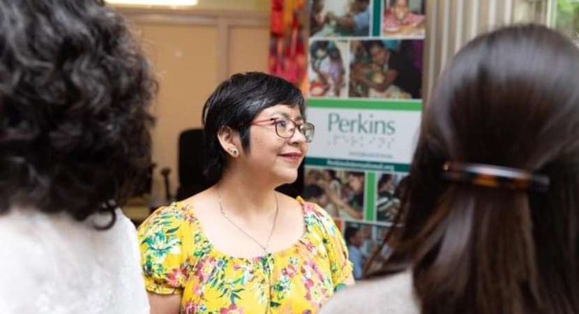 Una mujer de cabello oscuro y corto, vistiendo una camisa amarilla estampada, en el centro de la imagen mirando hacia su izquierda. Al fondo, un póster de Perkins.