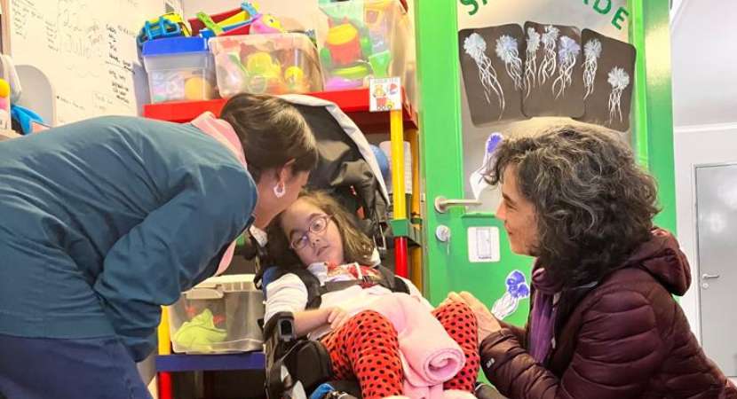 En un salón de clases colorido, una niña reclinada en su silla de ruedas encuantos dos maestras se abaixam a sua altura ´´ara fazer contato visual