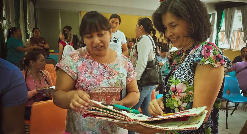 Dos mujeres lado a lado en un salón mirando un libro y sonriendo. En el fondo, varias mujeres están sentadas y de pie charlando entre ellas.