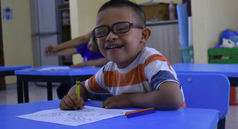 Un niño está sentado en su pupitre de clase. Lleva una camiseta colorida a rayas en azul, amarillo y naranja. Sonríe a la cámara mientras sostiene un crayón amarillo con el que está dibujando en un papel en su escritorio