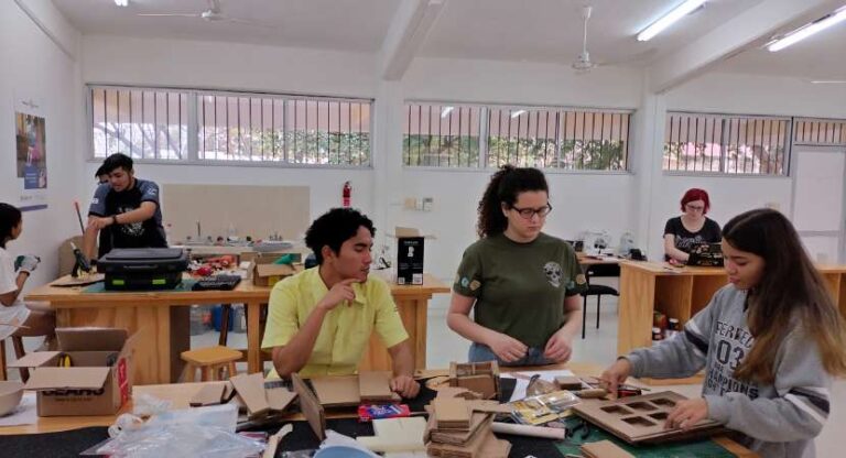 Un joven y dos jóvenes mujeres están alrededor de una mesa de trabajo llena de cartón, herramientas de oficina y diseño, y otros materiales. Hay otras dos mesas de trabajo detrás de ellos donde también hay una persona trabajando.