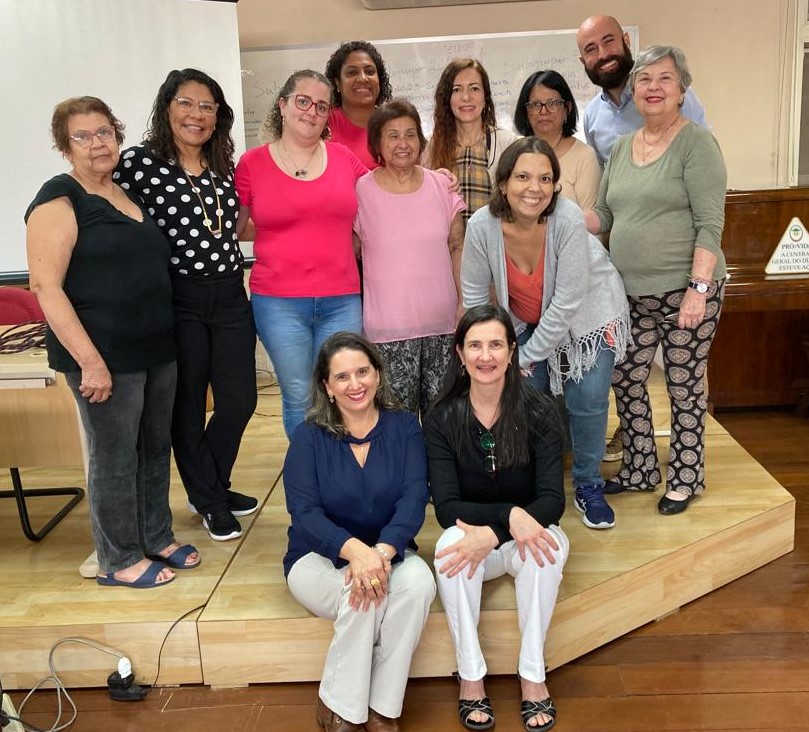 Foto da equipo do grupo estrategico sentido Perkins Brasil composto por 12 pessoas entre homens e mulhers. 10 pessoas estão de pé em um pequeno palco. Duas mulheres sentadas a frente do grupo.