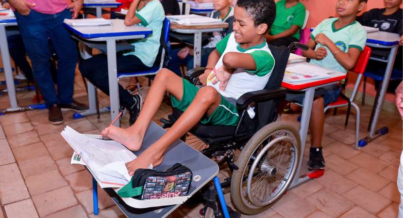 
Em um ambiente de sala de aula, um aluno está sentado em uma cadeira de rodas adaptada com uma prancha inclinada segurando seu caderno e escrevendo com um lápis usando o pé. Ao fundo, outros alunos sem deficiências estão estudando em carteiras.