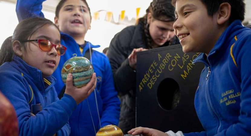 Foto de un grupo de tres estudiantes vestidos con uniforme azul. La niña con gafas sostiene un objeto redondo y colorido hecho de arte y manualidades. Un niño sonríe y otro sostiene una cuerda.