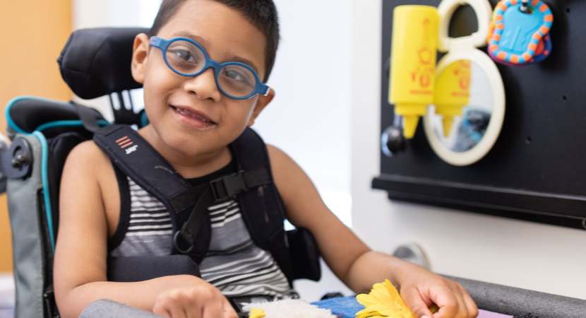 Un niño con gafas azules sentado en una silla de ruedas. Frente a él, hay una mesa con algunos juguetes coloridos. Él mira a la cámara y sonríe.
