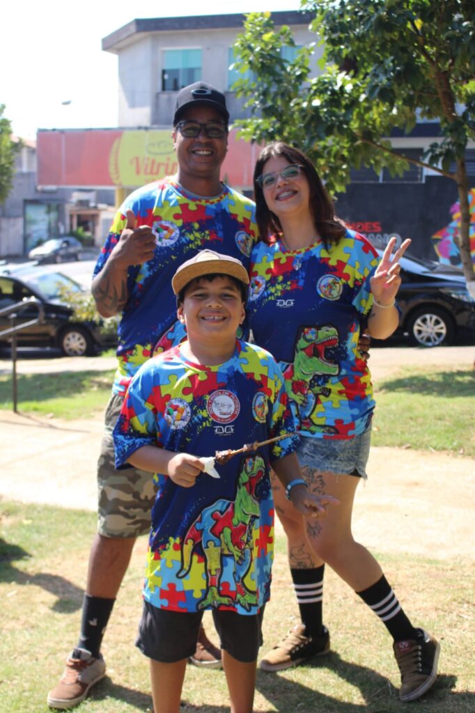 De pé e abraçados Thaís e seu marido Marcio, sorriem para a camera. À frente deles, seu filho Castiel, também sorri. Os três vestem camisetas coloridas que representam a causa do autismo.