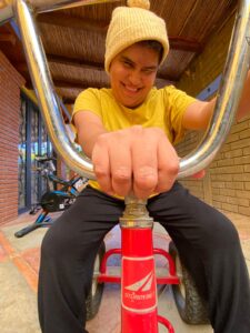 Una chica posa para una foto sentada en un triciclo rojo, agarrando el manillar con una mano. Lleva una camiseta y gorra amarilla.