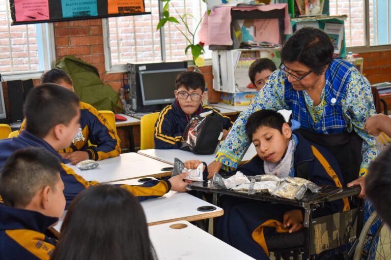 Siete alumnos se sientan en pupitres enfrentados a un alumno en silla de ruedas con su profesor detrás. El alumno en silla de ruedas pasa un objeto al alumno que tiene delante.