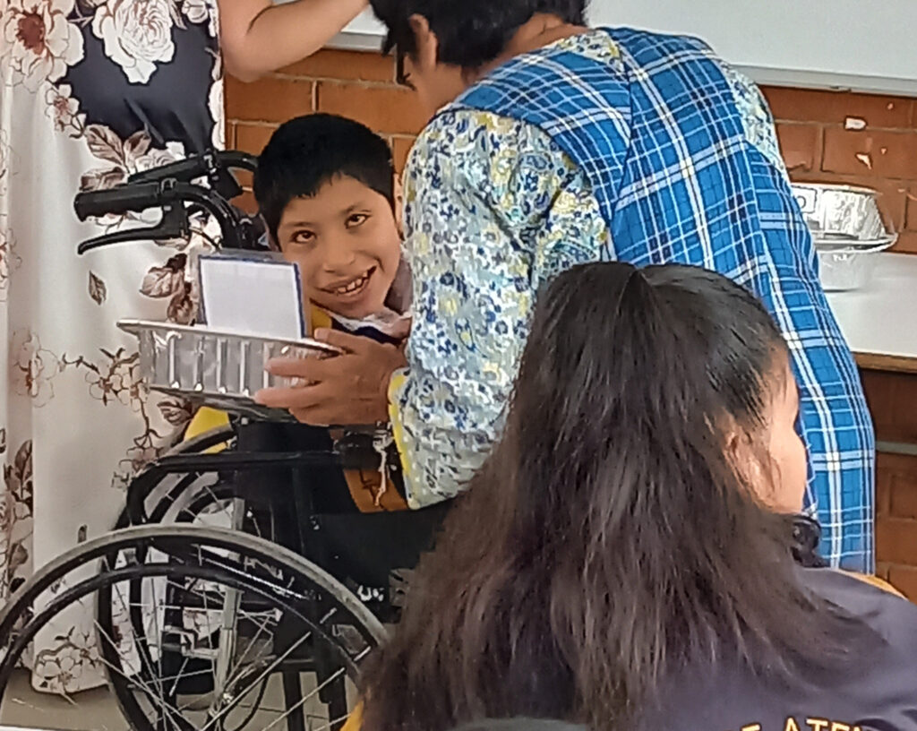 Un alumno mira a su profesora, que lleva un chaleco azul. Están realizando una actividad con una hoja de papel y una sartén metálica.